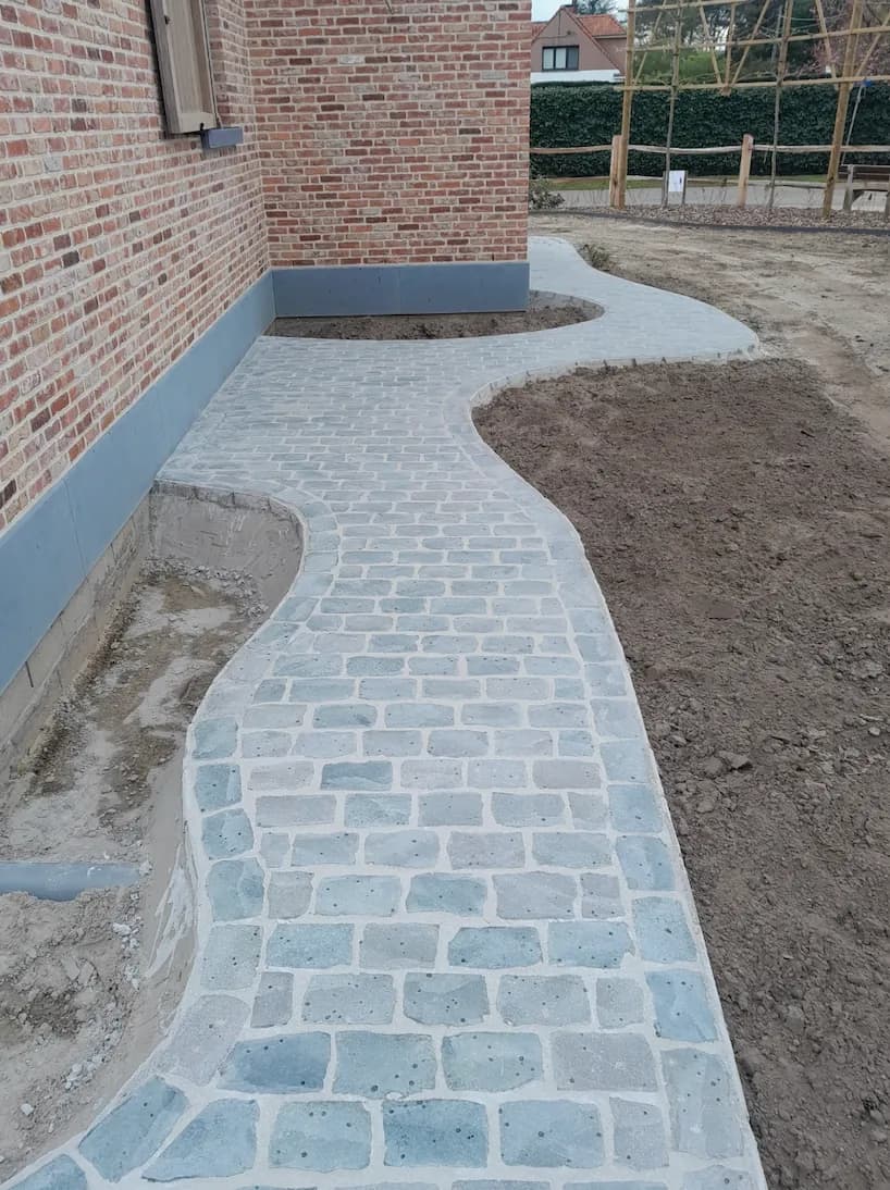 Slingerend tuinpad in lichte kasseien langs een bakstenen gevel, met tuin in aanleg